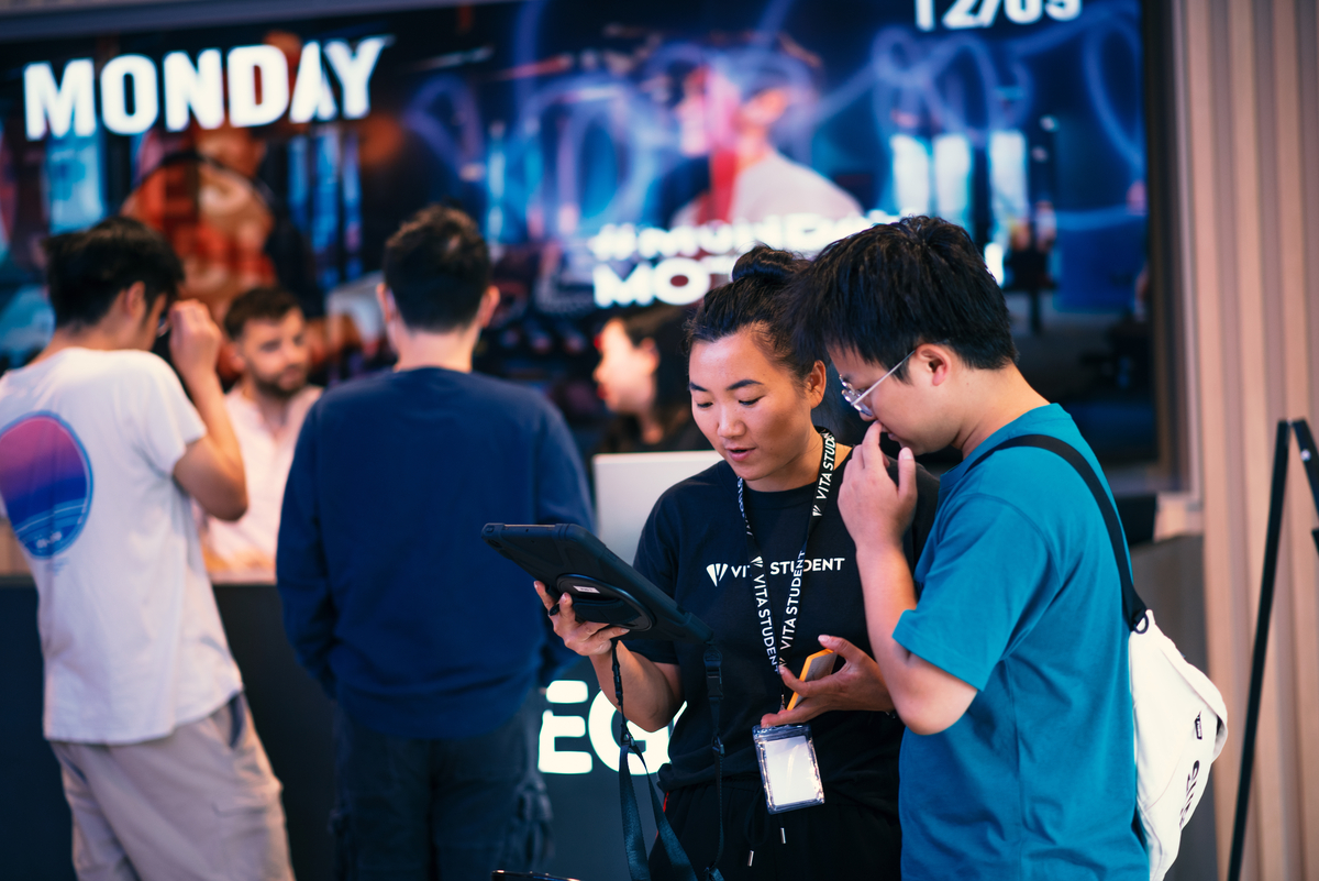 Two people stand in the foreground, looking at a tablet. One wears a student lanyard and a black shirt. In the background, several others are gathered in front of a screen displaying the word "MONDAY" with a date.